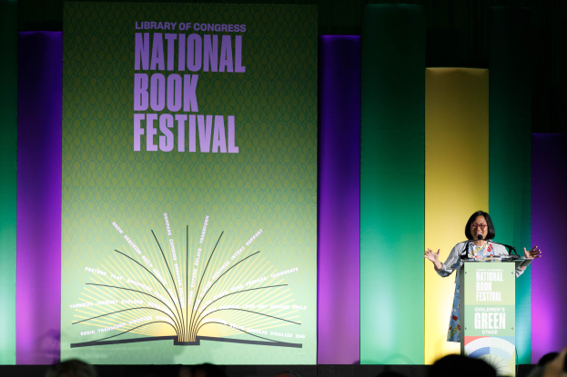 Eine Frau steht vor einem Publikum an einem Rednerpult und spricht in ein Mikrofon, hinter ihr ist eine Tafel mit Text zu sehen.