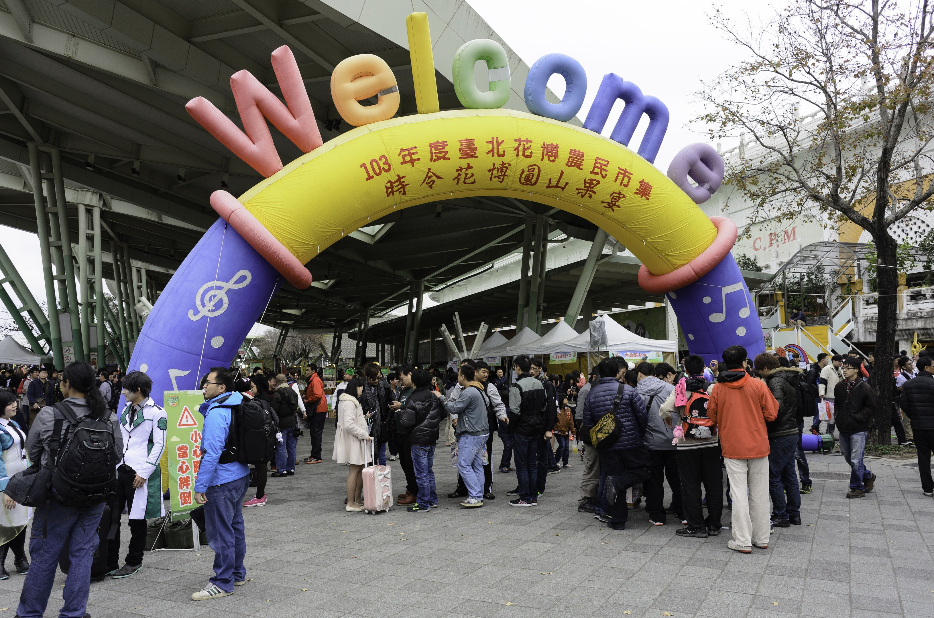 Eine Gruppe von Menschen steht vor einem Willkommensbogen bei einem Outdoor-Event, mit Zelten, Bäumen und Gebäuden im Hintergrund unter einem klaren blauen Himmel, einige tragen Taschen.