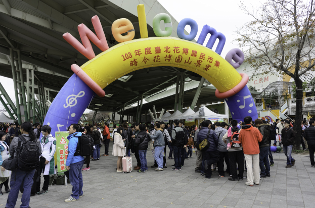 Eine Gruppe von Menschen steht vor einem Willkommensbogen bei einem Outdoor-Event, mit Zelten, Bäumen und Gebäuden im Hintergrund unter einem klaren blauen Himmel, einige tragen Taschen.