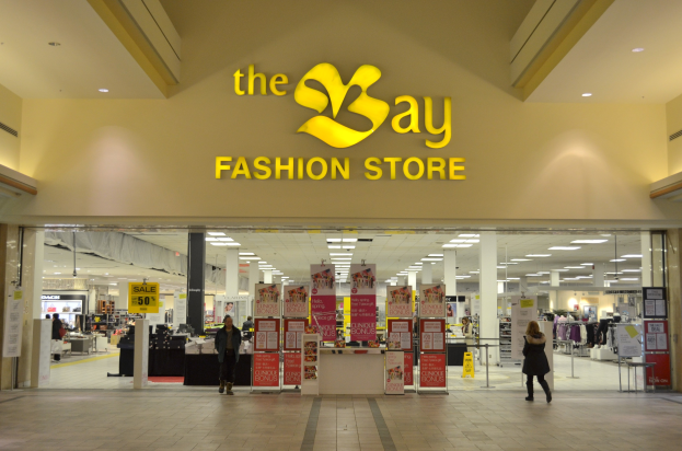 Außenansicht eines Bay Fashion-Geschäfts in einem Einkaufszentrum mit Passanten, Wandtexttafeln, Säulen, Deckenleuchten und Hintergrundauslagen.