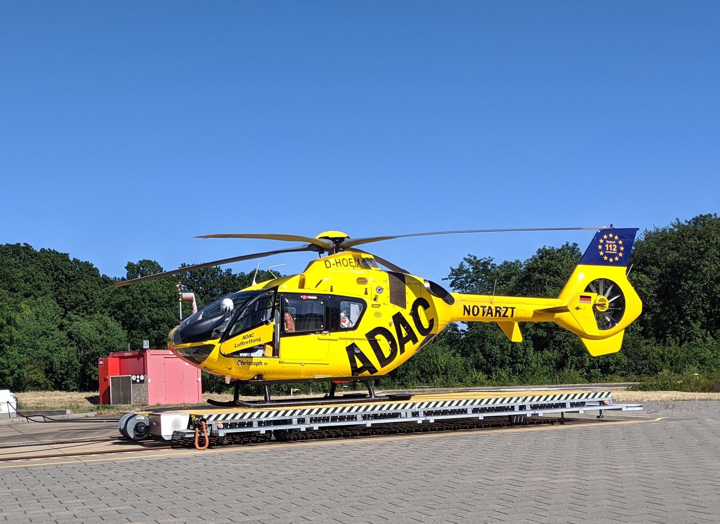 Gelbes Hubschrauber auf einem Flughafenvorfeld mit Bäumen und einem klaren blauen Himmel, umgeben von Gegenständen, die wahrscheinlich mit Luftrettungsdiensten in Deutschland in Zusammenhang stehen.