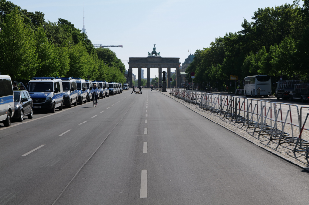 Lange Reihe von Polizeiwagen auf der Seite einer Straße vor dem Brandenburger Tor in Berlin, Deutschland, mit Menschen auf Fahrrädern und Barrieren auf der Straße, Bäumen an den Seiten, einem Bogen mit Statuen im Hintergrund und dem Himmel darüber.