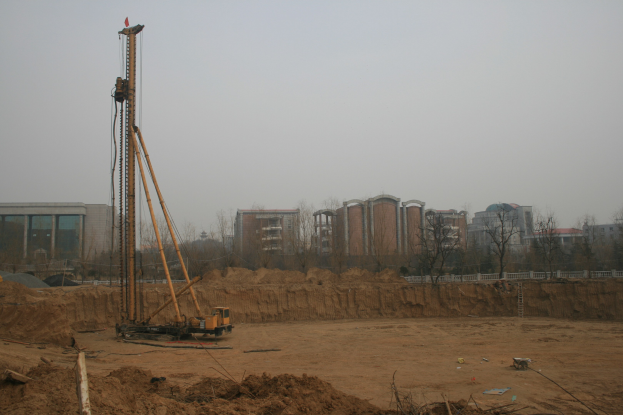Eine Baustelle mit einem Kran in der Mitte, umgeben von Gebäuden, Bäumen und einem klaren blauen Himmel, mit bodenbedecktem Grund und verstreuten Gegenständen.