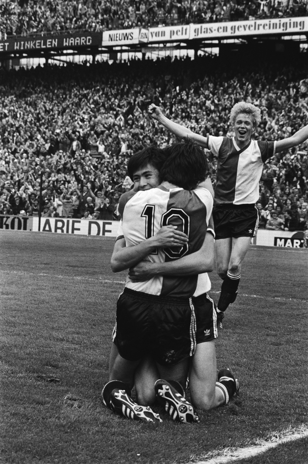 Drei Fußballspieler feiern ein Tor auf dem Boden vor einer jubelnden Menge in einer schwarz-weißen Stadionszene.