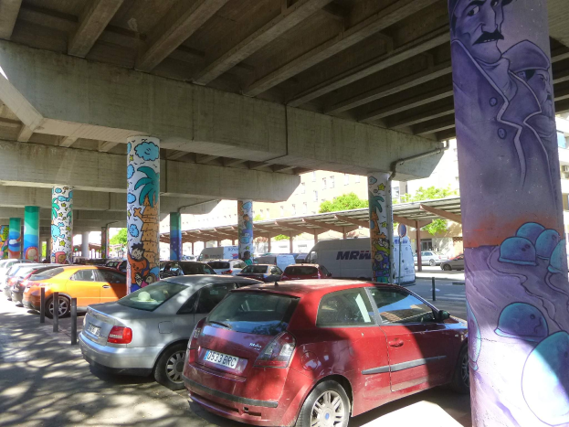 Ein Parkplatz mit zahlreichen geparkten Autos unter einer Brücke, mit Graffiti an seinen Pfeilern und nächsten Gebäuden, mit Bäumen im Hintergrund und einem Van, der unter den Fahrzeugen geparkt ist.