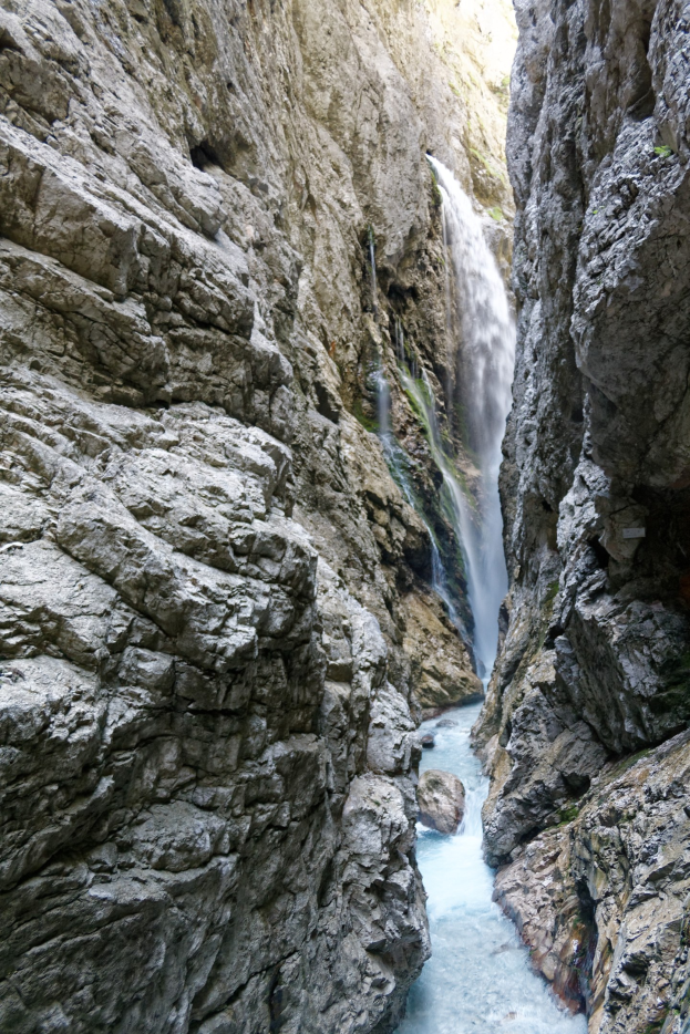 Ein kleiner Wasserfall stürzt über zerklüftete Felsen in einem steinigen Tal hinab, umgeben von saftig grünen Hügeln unter strahlendem Sonnenschein.