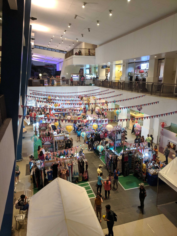 Ein volles Einkaufszentrum mit Ständen, hängenden Kleidern, Dekorationsfahnen, Ballons, sitzenden und stehenden Menschen, Geländern, Säulen und Deckenleuchten, ähnlich einem Weihnachtsmarkt im Mall of Asia.