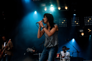 Eine Frau singt auf der Bühne in ein Mikrofon, flankiert von einem Gitarristen links und einem Schlagzeuger im Hintergrund, alles in helles Bühnenlicht getaucht.