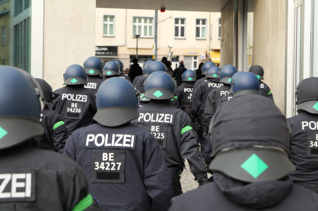 Gruppe von Polizisten in Uniform und Helmen vor einem Gebäude stehend, mit Menschen, Pfählen und anderen Gebäuden im Hintergrund.