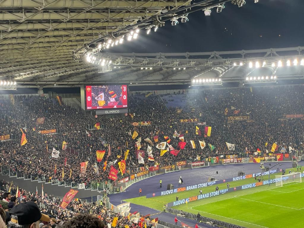 Eine große Menge in einem Stadion schwenkt Fahnen und Banner, während sie auf Gras sitzt, mit einem Bildschirm im Hintergrund und hellen Scheinwerfern, die die aufgeregte Stimmung erleuchten, bevor ein Fußballspiel beginnt.