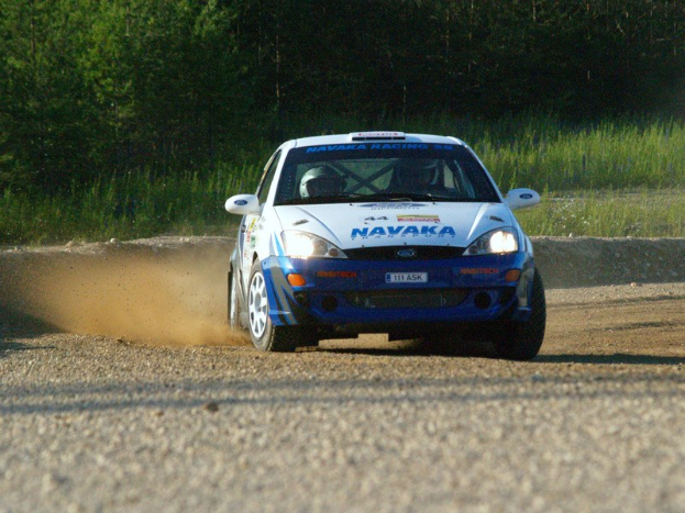 Blauer und weißer Rallye-Wagen mit zwei Helmträgern auf einer Schotterstraße, umgeben von Vegetation.