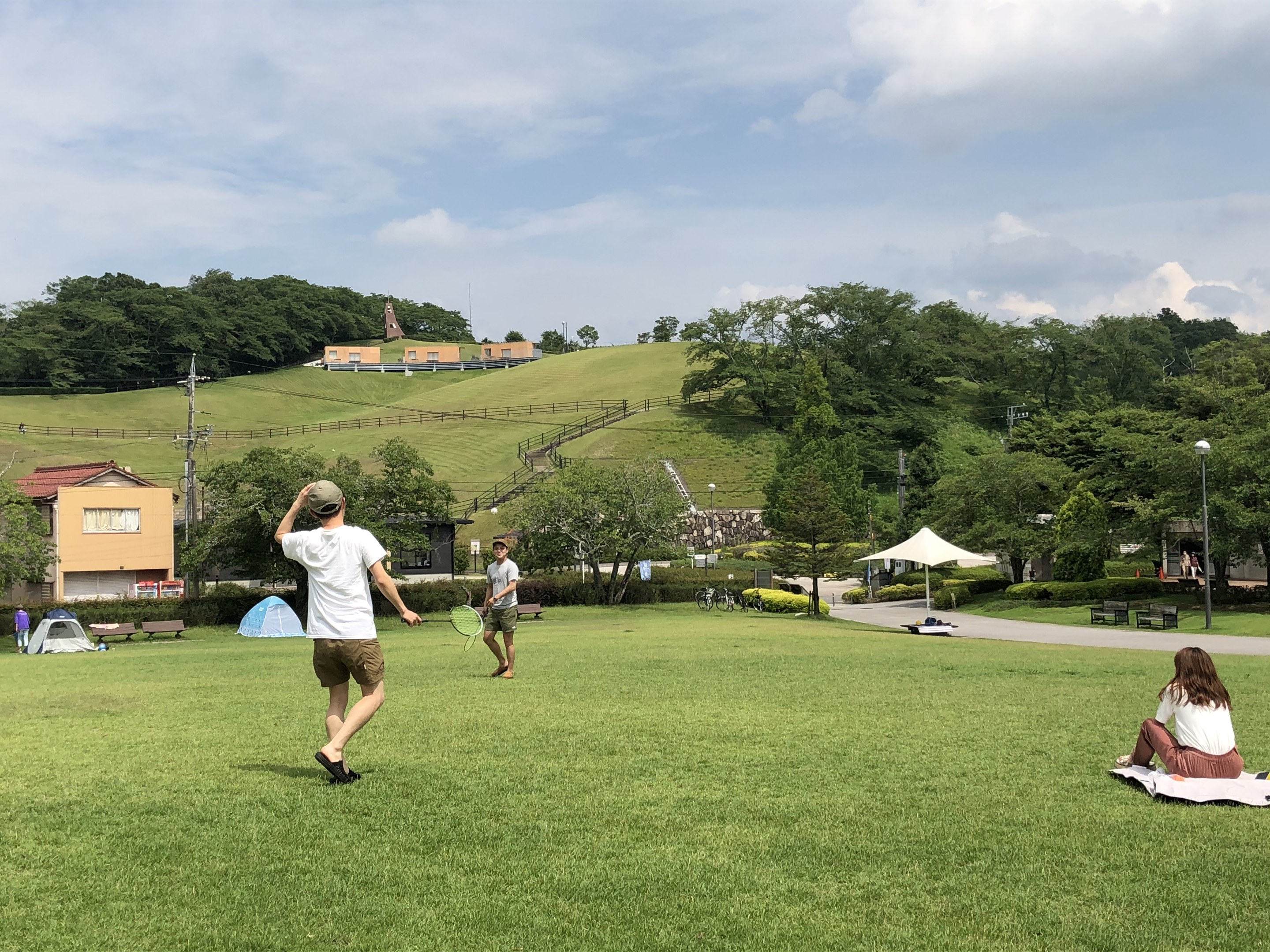 Gruppe von Menschen, die Badminton in einem Park spielt, mit einem Mann, der einen Schläger hält und auf einer Decke auf dem Gras sitzt, mit Zelten, Gebäuden, Hügeln und einem bewölkten Himmel im Hintergrund.