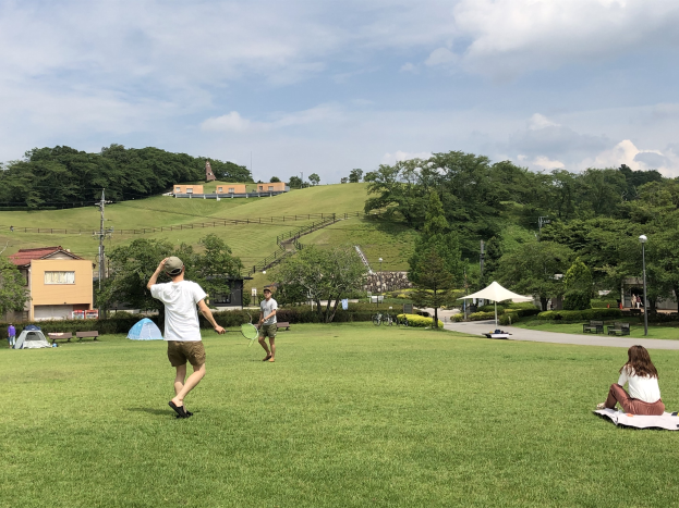 Gruppe von Menschen, die Badminton in einem Park spielt, mit einem Mann, der einen Schläger hält und auf einer Decke auf dem Gras sitzt, mit Zelten, Gebäuden, Hügeln und einem bewölkten Himmel im Hintergrund.
