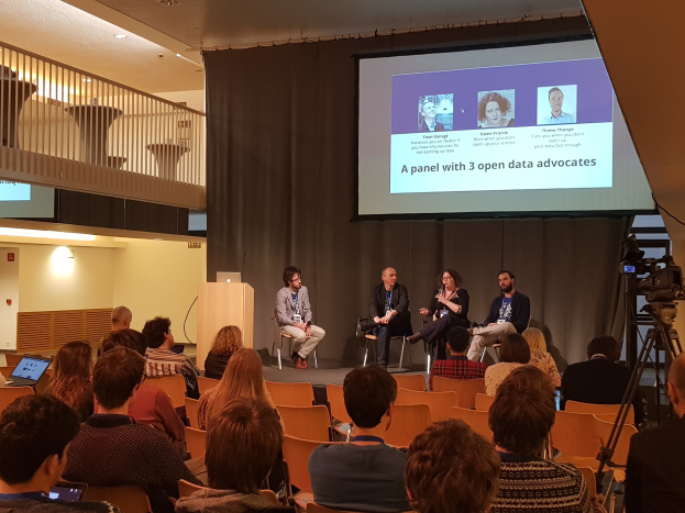 Eine Gruppe von Menschen sitzt auf Stühlen vor einem Projektionsschirm mit einem Laptop auf einem Tisch, eine Kamera auf einem Stativ rechts daneben und Geländern, Lampen und einer Wand im Hintergrund, die an einer Podiumsdiskussion über Open-Data-Aktivisten teilnehmen.
