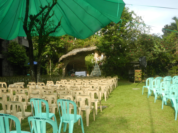 Eine Hochzeitszeremonie im Garten mit grünen Stühlen und Schirmen, umgeben von üppiger Vegetation, mit einem Gebäude und einem Schuppen im Hintergrund.