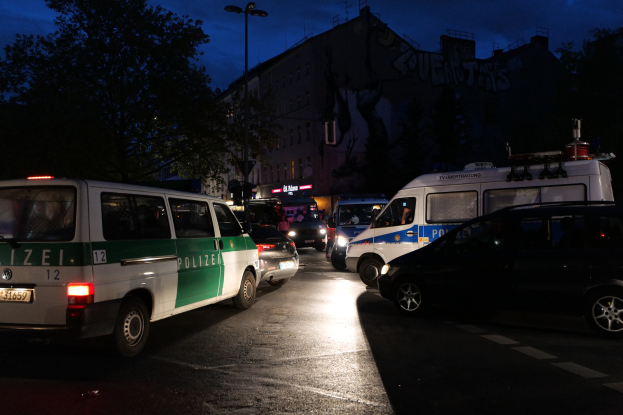 Eine Gruppe von Polizeiwagen, darunter ein Van, ein Auto und ein Rad, parken nachts am Straßenrand mit Gebäuden, Bäumen, Laternenmasten und einem sternenklaren Himmel im Hintergrund.