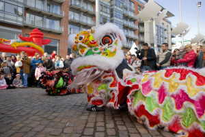 Ein lebendiges chinesisches Neujahrsfest in Amsterdam mit einem Löwen tanzen im Vordergrund und einer Zuschauermenge, einige halten Kameras, vor einem Hintergrund aus Gebäuden, Laternenmasten und einem klaren blauen Himmel.