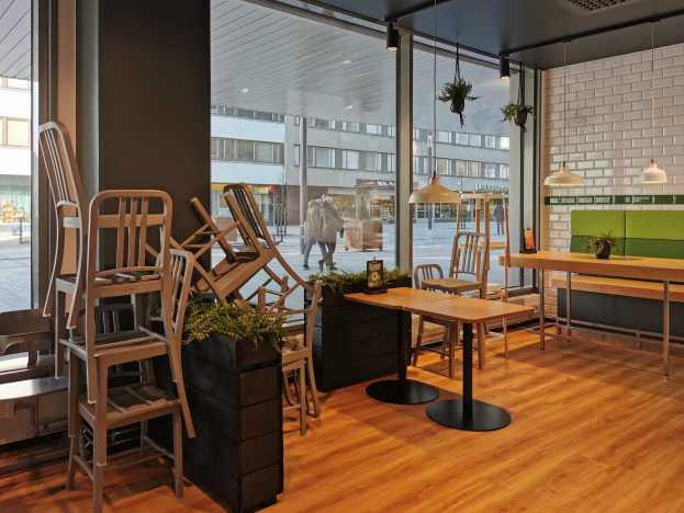 Ein Restaurantinnenraum mit Holztischen und -stühlen, die durch ein großes Fenster zu sehen sind, das eine Außenansicht von Berlin mit Passanten, Gebäuden und Straßenelementen zeigt.