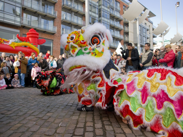 Ein lebendiges chinesisches Neujahrsfest in Amsterdam mit einer Löwen-Tanzvorstellung vor einer Zuschauermenge, die Kameras hält, vor einer Kulisse aus Gebäuden, Laternenmasten und einem klaren blauen Himmel.