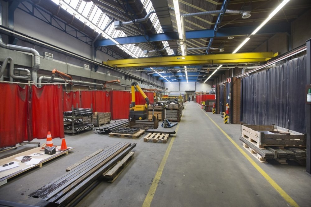 Großer Lagerraum mit Holzpaletten, Metallobjekten, Verkehrskegeln, Maschinenfahrzeug, rotem Vorhang, Deckenrohren, Lampen und einer verhängten Wand auf der rechten Seite.