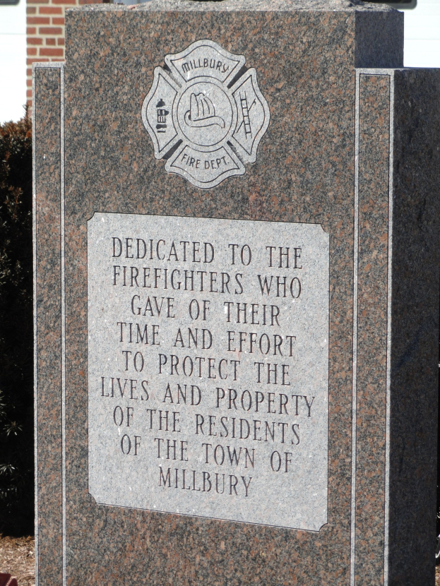 Gedenkstein mit einer Plakette für Feuerwehrleute in Millbury, vor einem Backsteingebäude mit umliegenden Pflanzen und Bäumen aufgestellt.