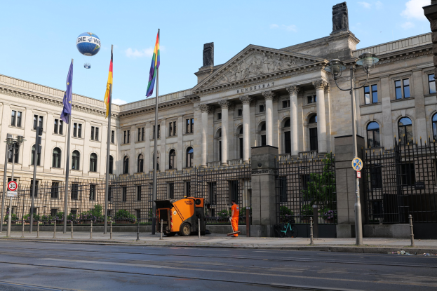 Großes beige Gebäude mit vielen Fenstern, identifiziert als Bundestag in Berlin, Deutschland, umgeben von Straßeninfrastruktur, Füüballern, Fahrzeugen und Bäumen unter einem bewölktem Himmel.