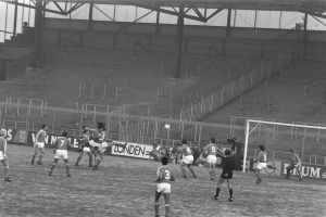 Eine Gruppe von Männern, die Füßball auf einem saftig grünen Feld in einem Stadion spielen, mit einem Torpfosten auf der rechten Seite, Treppen und Geländern im Hintergrund und Bannern mit Text, dargestellt in Schwarz-Weiß.
