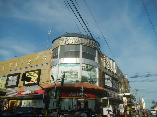 Eine belebte Stadtstraße mit fahrenden Fahrzeugen, Fußgängern auf dem Gehweg und dem Royal Mall-Einkaufszentrum im Hintergrund, umgeben von Bäumen, Versorgungsmasten und Drähten unter einem Himmel.