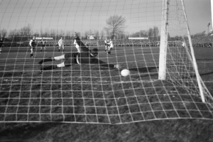 Schwarzes und weißes Foto eines Fußballtorwarts beim Abwehrversuch, während andere Spieler das Spiel auf dem Feld fortsetzen, mit Bäumen, Pfosten und Werbetafeln im Hintergrund bei klarem Himmel.