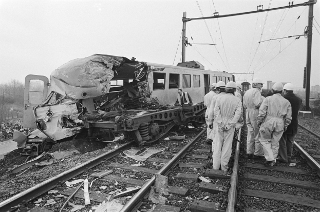 Ein Schwarz-Weiß-Bild einer Zugkollision auf den Schienen mit Einsatzkräften in Helmen und einem Hintergrund aus Strommästen, Bäumen, Gebäuden und einem klaren Himmel.