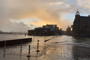 ├ťberflutete Stra├če in Hamburg, Deutschland, mit Wasser bedecktem Asphalt, Masten, Schildern, Geb├Ąuden mit Fenstern, einer Br├╝cke und einem bew├Âlktem Himmel im Hintergrund.
