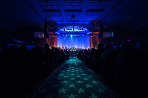 Ein großer Auditorium mit Sitzenden, eine Bühne mit Musikern und eine Decke mit Lichtern und Fernsehgeräten, was auf einen Konzertsaal für die Nationale Convention 2019 hinweist.