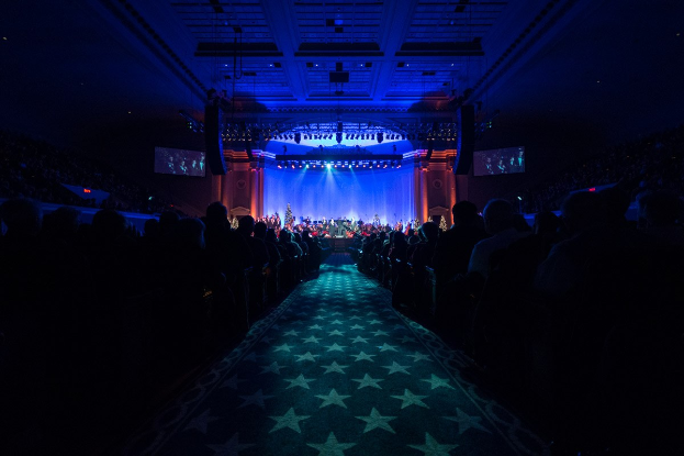 Ein großer Auditorium mit Sitzenden, eine Bühne mit Musikern und eine Decke mit Lichtern und Fernsehgeräten, was auf einen Konzertsaal für die Nationale Convention 2019 hinweist.