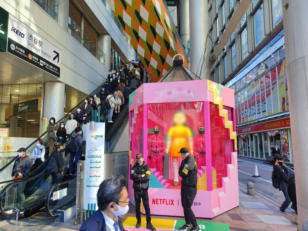 Eine Gruppe von Menschen steht vor einem Glaswand-Gebäude mit Säulen und Schildern, einige tragen Masken, während eine Rolltreppe mit Menschen zu sehen ist. In der Mitte steht eine pinkfarbene Box mit Schaufensterpuppen und der Aufschrift "Netflix's new Netflix booth in Tokyo".