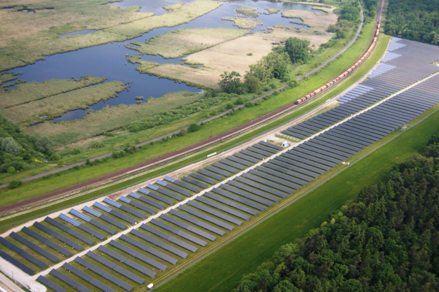 Luftaufnahme eines Solarparks mit Panelen, umgeben von Bäumen, Gras, Wasser und einer nahen Bahntrasse.