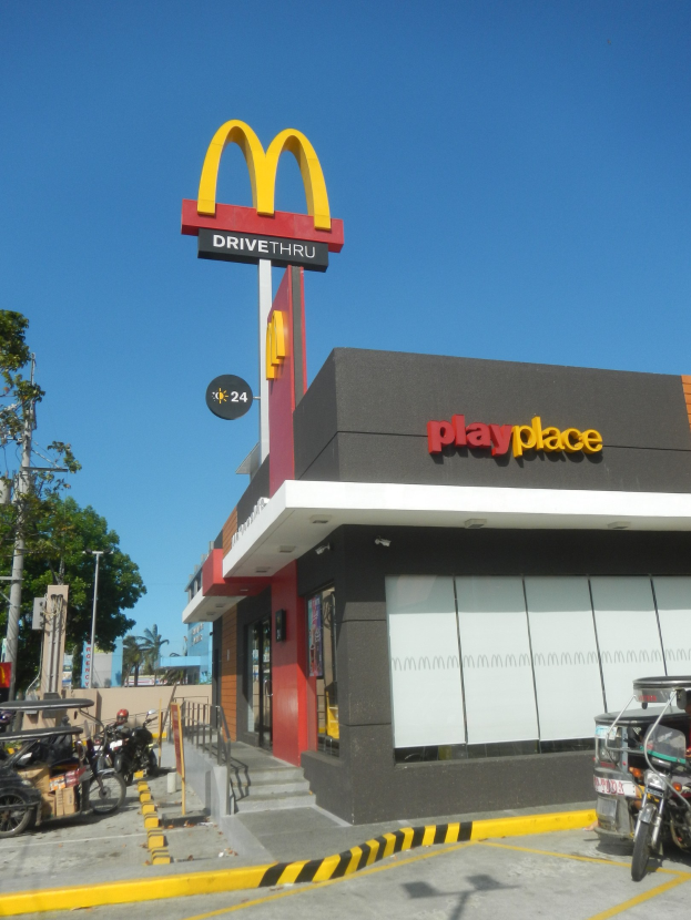 Ein McDonald's-Restaurant mit einem "Playplace"-Schild, umgeben von Fahrzeugen, Bäumen, Polen und einem klaren blauen Himmel.