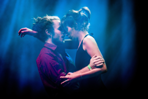 Zwei Menschen umarmen und küssen sich auf einer Bühne mit Hintergrundbeleuchtung.