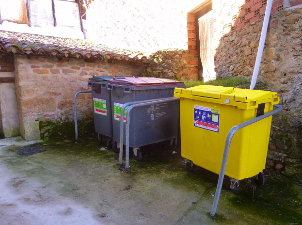 Zwei gelbe und graue Recyclingcontainer auf Rädern nebeneinander vor einem Gebäude mit einer Wand und einem Fenster.