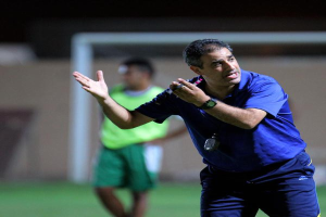 Ein Mann in einem blauen T-Shirt und Hose steht auf einem Fußballfeld und hält ein Mikrofon in der Hand, während er zu seinen Spielern gestikuliert. Im Hintergrund ist eine Person in einem grünen Kleid, ein Tor und eine Wand zu sehen.