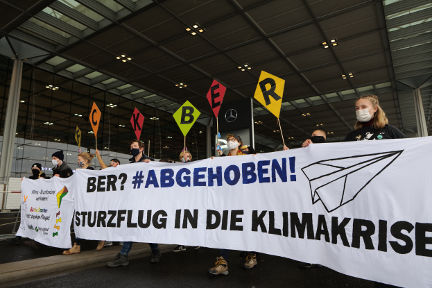 Eine Gruppe maskierter Individuals mit einem Banner "Sturzflug in die Klimakrise" steht vor einem gläsernen Gebäude mit Säulen, beleuchtet von Deckenlampen.