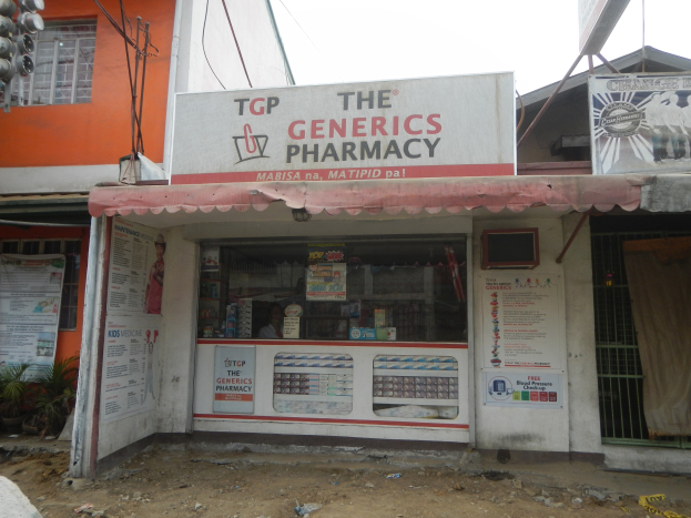 Apotheken-Schriftzug "The Generics Pharmacy" in städtischer Umgebung mit Gebäuden, Fahrzeugen und einem klaren Himmel.