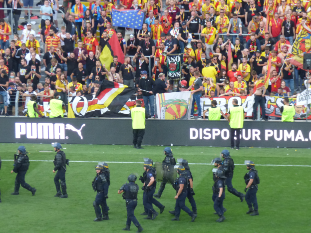 Polizeibeamte in Helmen und taktischer Ausrüstung überqueren ein Fußballfeld und tragen Waffen, während Zuschauer Fahnen und Banner schwenken.