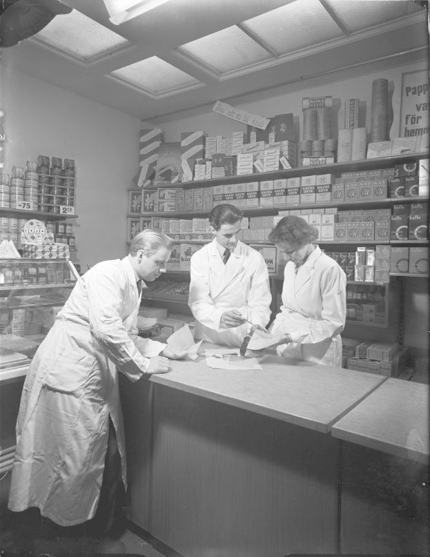 Drei Männer in weißen Kitteln an einem Apothekenschalter mit Papieren und Gegenständen, vor Regalen mit Artikeln, in Schwarz-Weiß.