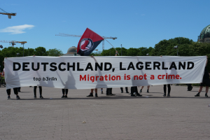 Eine Gruppe von Menschen hält ein Transparent mit der Aufschrift "Deutschland, Lagerland Migration ist kein Verbrechen" und eine Flagge, mit Straßeninfrastruktur, Bäumen, einem Gebäude und einem bewölkten Himmel im Hintergrund.