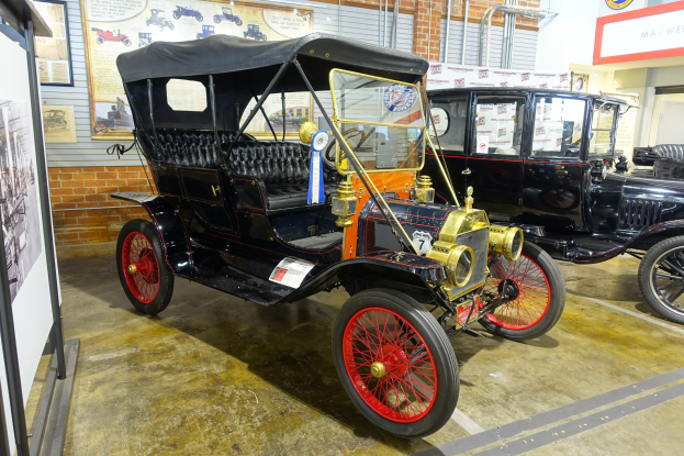 Ein Oldtimer in einem Museum ausgestellt mit Informationsschildern, Deckenlampen und sichtbaren Rohren, mit einem Rad im Fahrzeug.