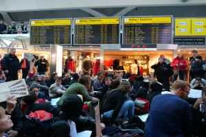 Eine große Gruppe von Menschen in einem Flughafen, einige sitzen mit Taschen und Papieren, andere stehen, mit Texttafeln, Schaufensterpuppen in Kleidern und Deckenlampen im Hintergrund, während einer Demonstration.