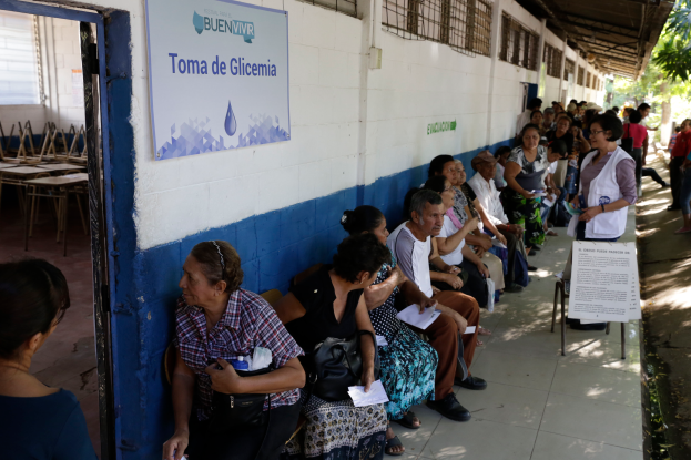 Eine Gruppe von Menschen sitzt auf Bänken vor einem Gebäude mit einer Texttafel an der Wand und einer Tür links, die an einer Wahllokalwarteschlange in Guadalajara, Mexiko, warten.