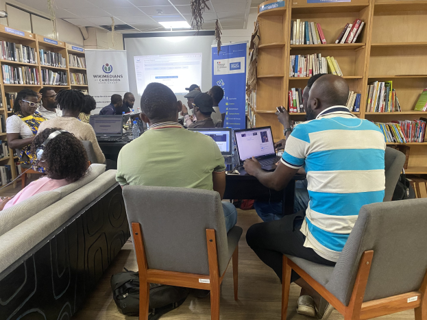 Eine Gruppe von Menschen sitzt an einem Tisch mit Laptops in einer Bibliothek, umgeben von Bücherregalen, mit einem Banner, Bildschirm und Deckenbeleuchtung im Hintergrund und Taschen auf dem Boden.