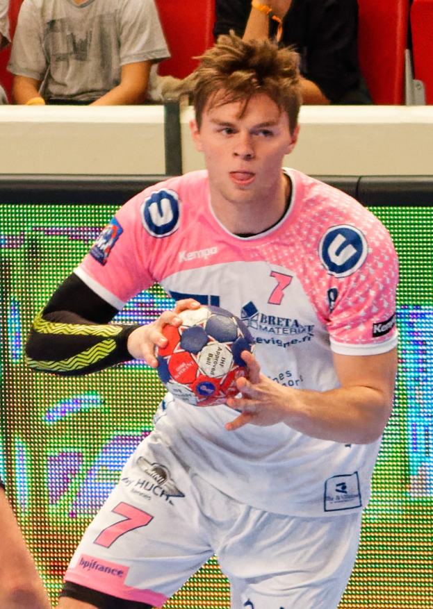 Ein Mann in einem pinken Hemd mit einem Fußball, daneben die Hand einer anderen Person und im Hintergrund sitzende Menschen.