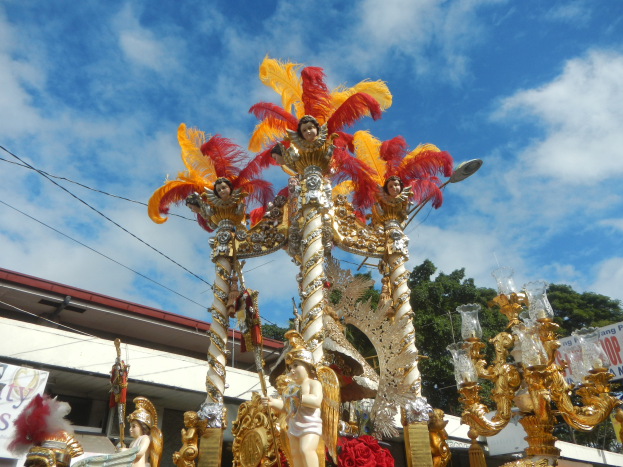 Ein Karnevalswagen mit goldenen und roten Federn, der Menschen trägt, ist in einem Umzug mit einem Gebäude, Bäumen, Drähten und einer Fahne in der Nähe zu sehen, unter einem sichtbaren Himmel.